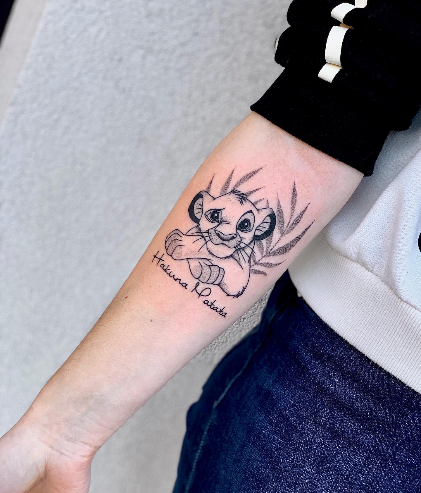 Forearm tattoo of a cartoon lion cub Simba with the text "Hakuna Matata" and a leaf background